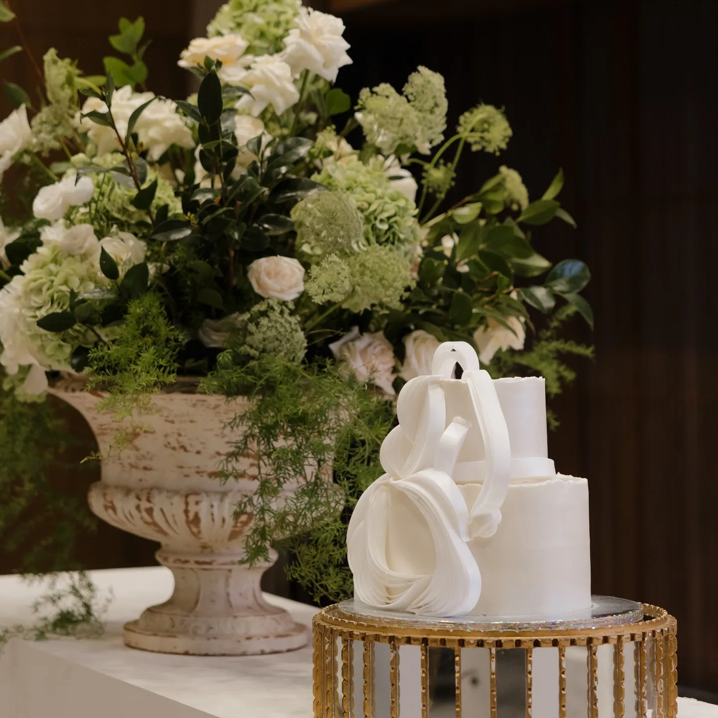 Pastel de bodas blanco sobre base dorada junto a una urna de piedra con flores
